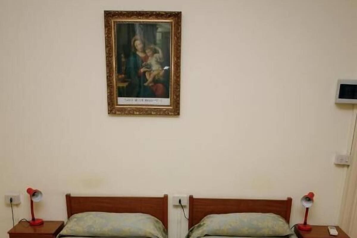 A double room with two single beds, each with a wooden headboard and a green floral bedspread. There are bedside tables with lamps, a framed picture on the wall, and a thermostat on the wall.