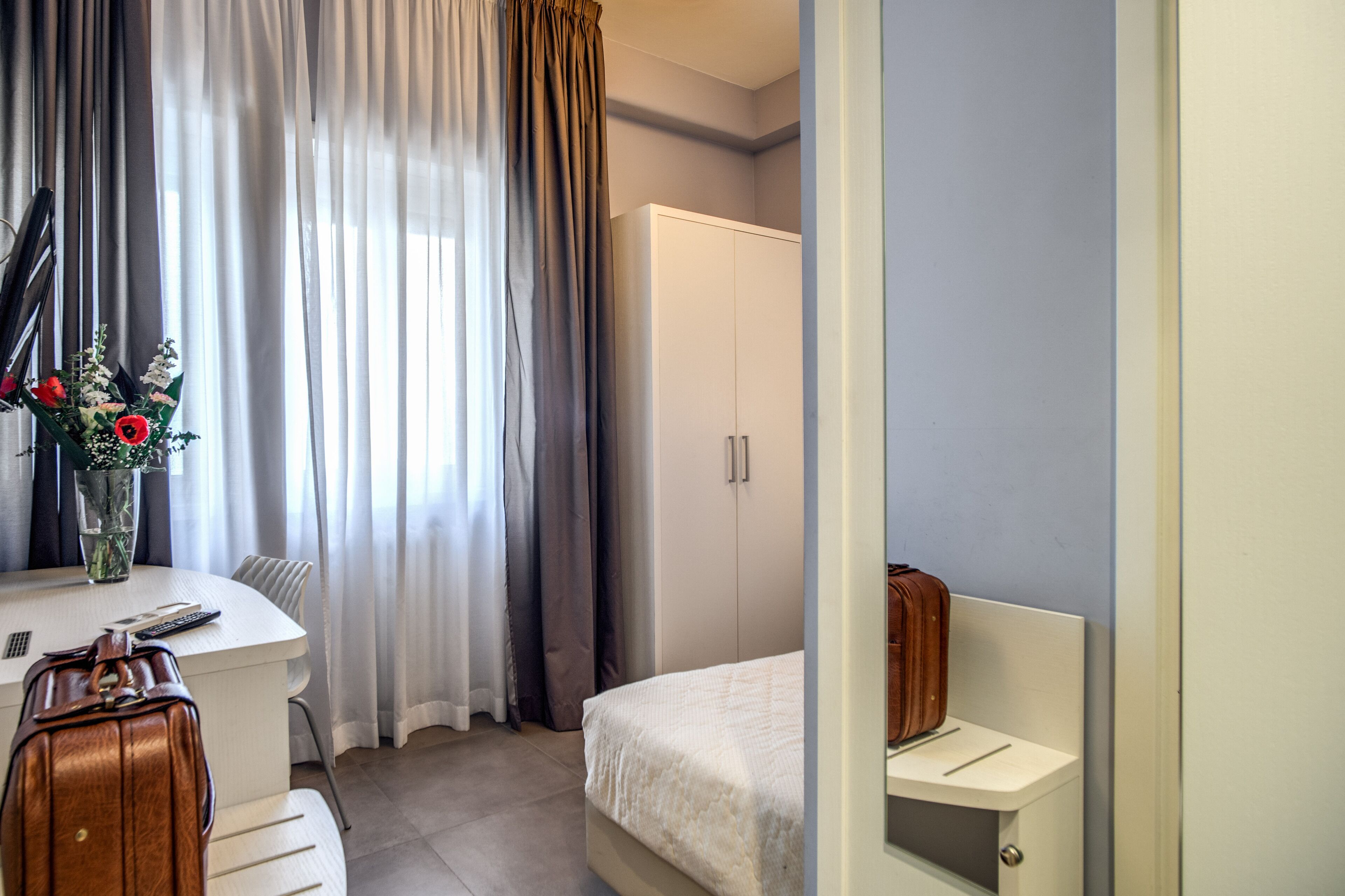 A hotel room with a bed, a desk with a chair, a white wardrobe, and a vase with flowers on the desk.