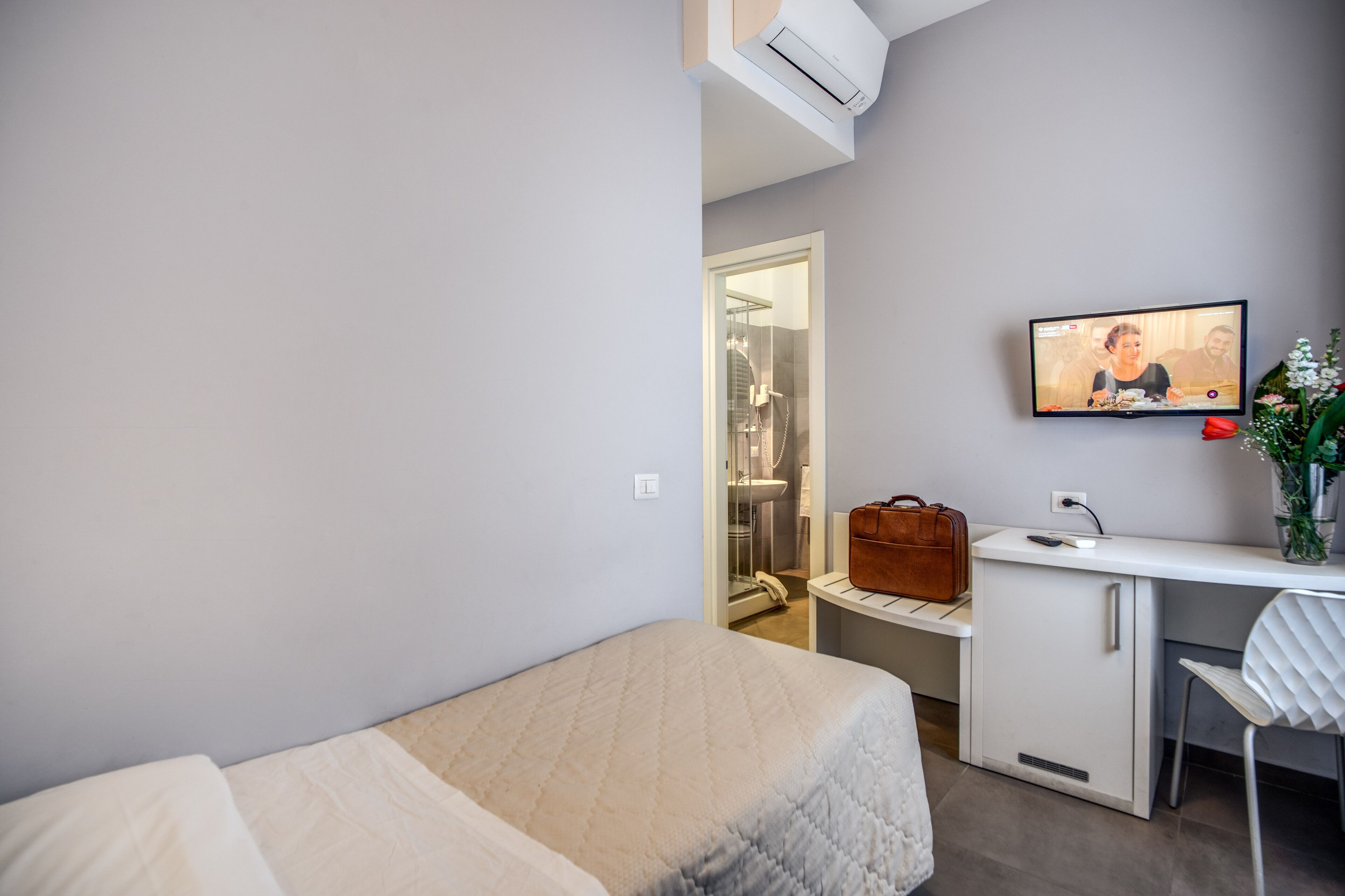 A modern hotel room with a bed, a desk with a computer, a small cabinet, and a shower area visible through an open door.