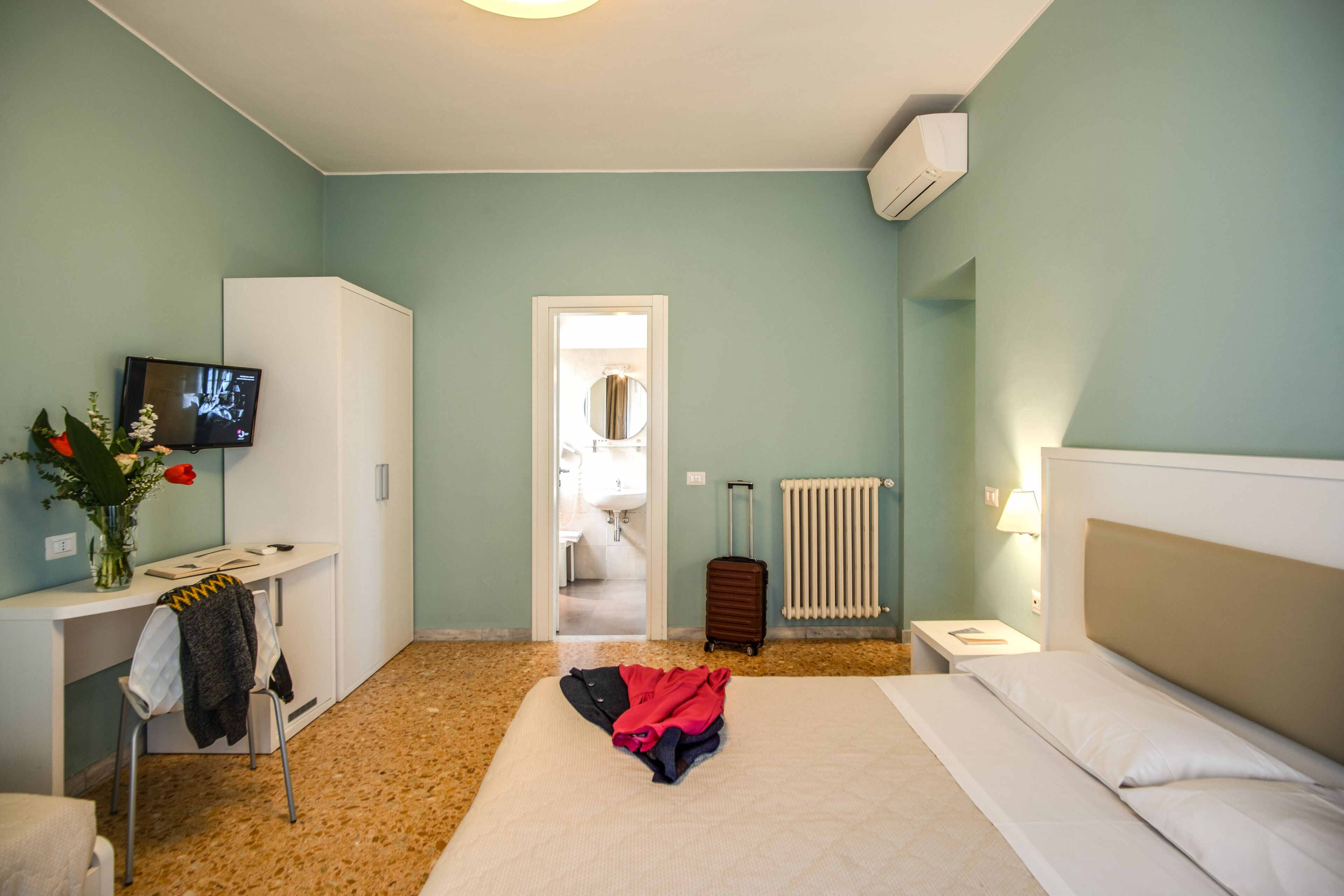 A hotel room with a bed, a desk with a TV, a radiator, and a suitcase.