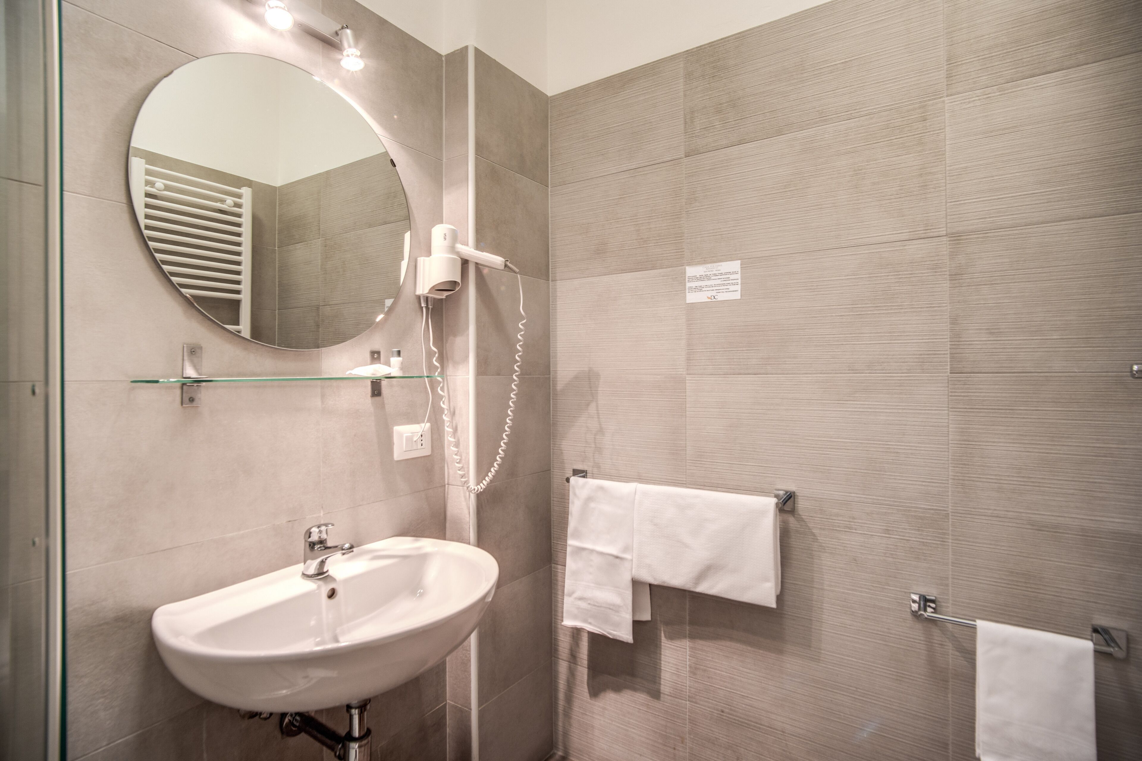 A modern bathroom with a large oval mirror, a white sink, and a glass shower enclosure.