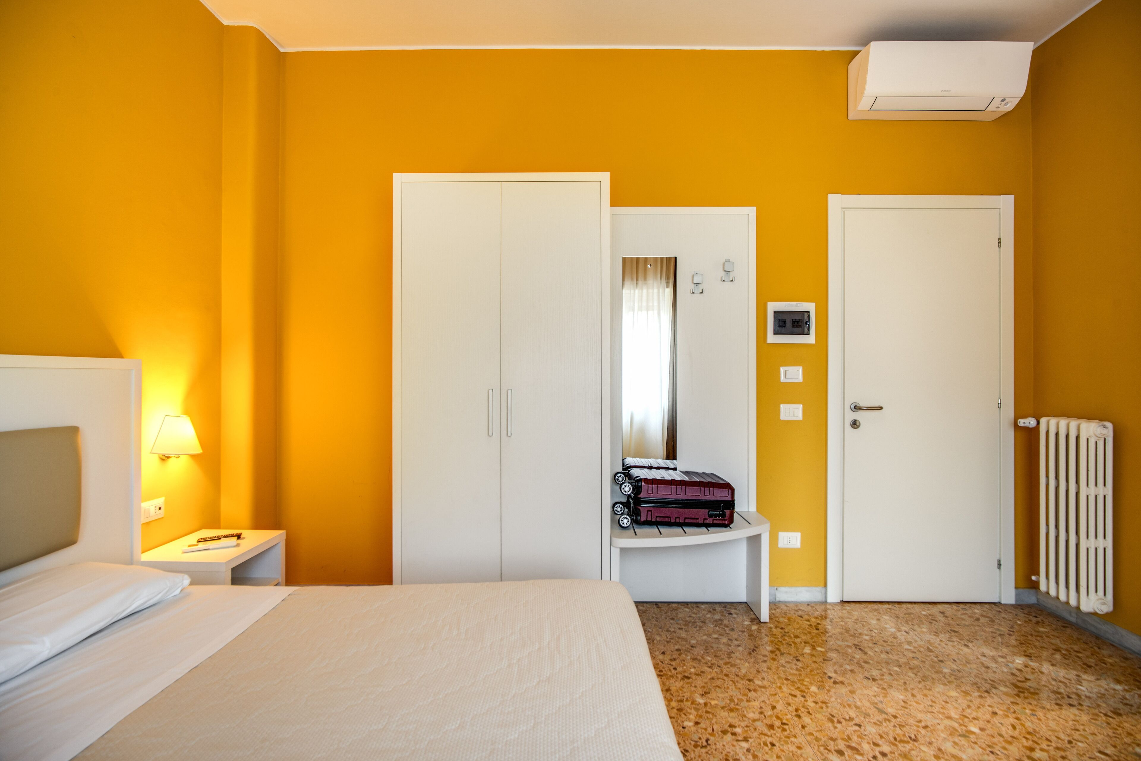 A hotel room with a bed, a nightstand, a wardrobe, a radiator, and a suitcase on a small table.