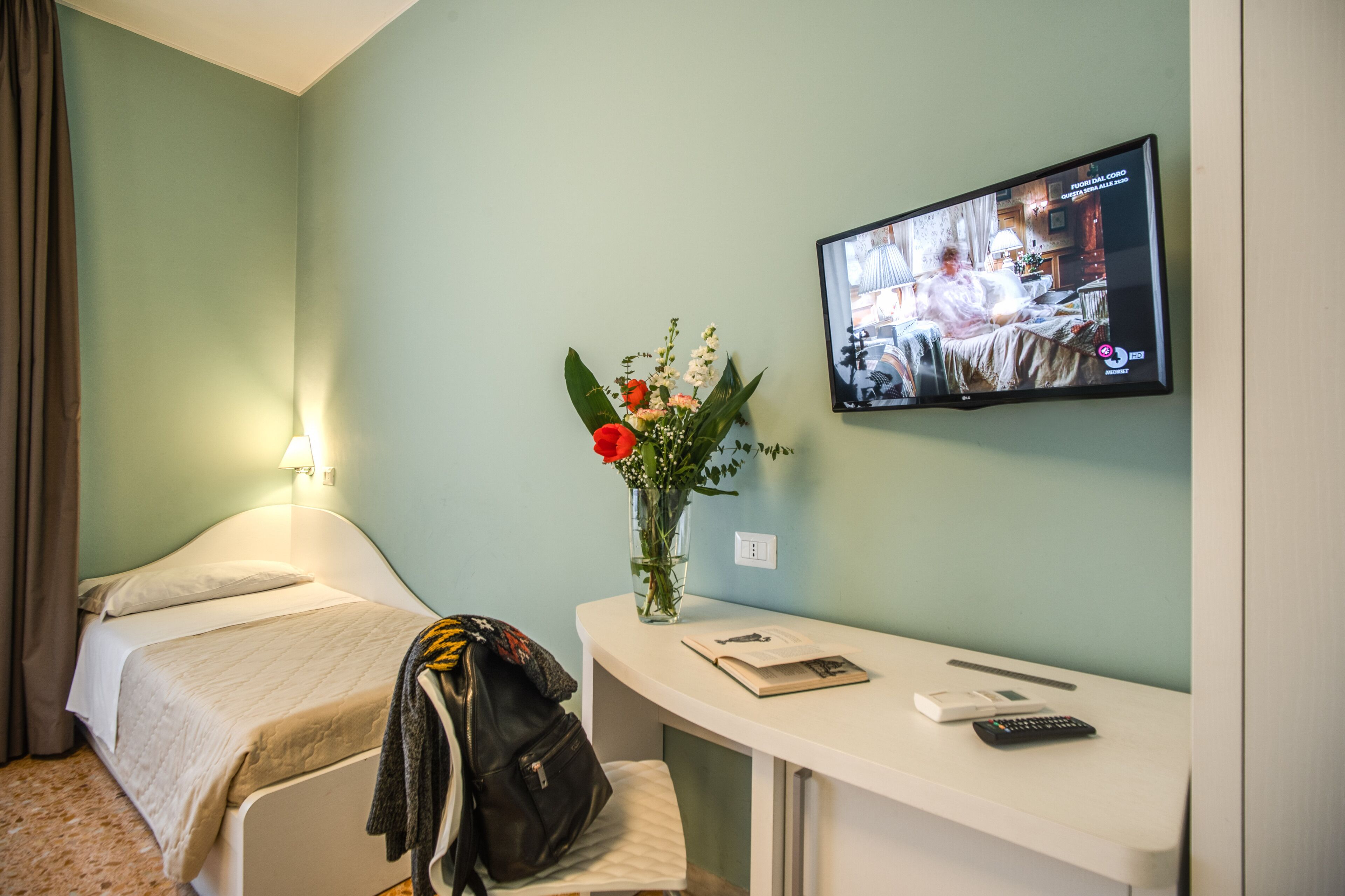A hotel room with a bed, a desk with a vase of flowers, a remote control, and a television displaying a news program.