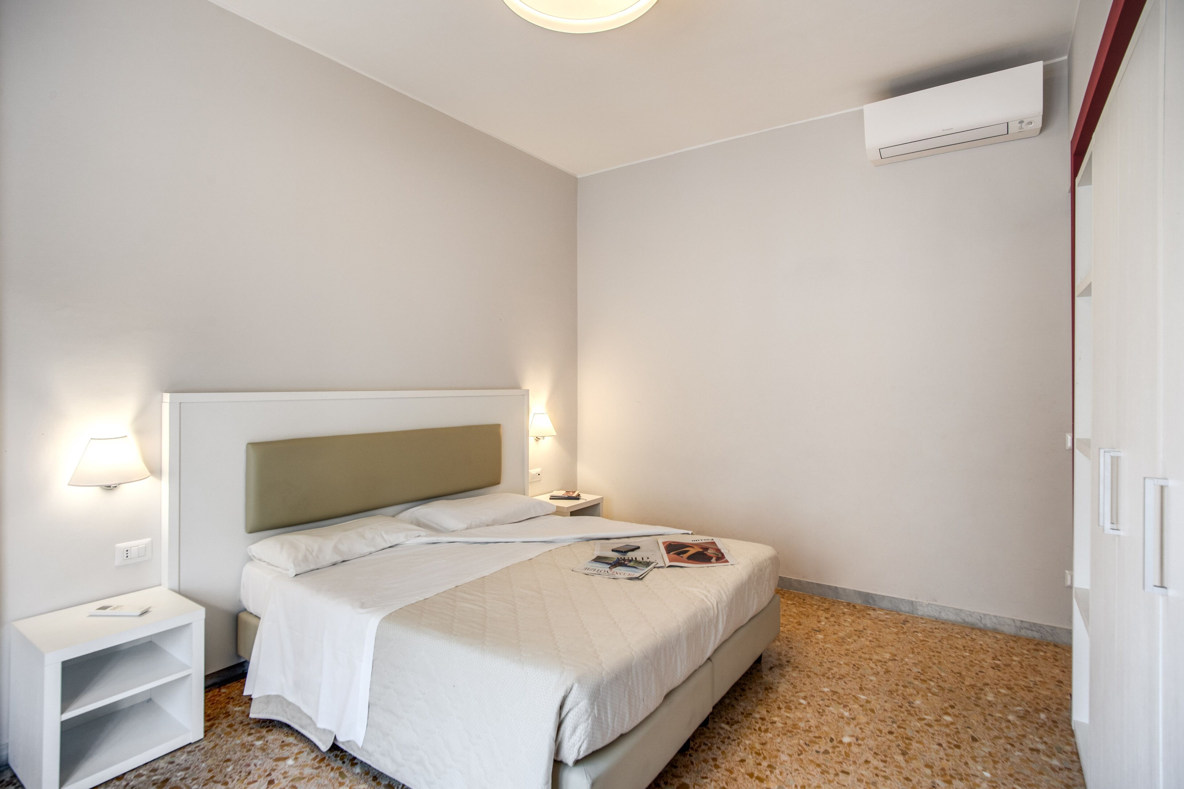 Une chambre d’hôtel comprenant un lit, des tables de chevet, une petite table, un climatiseur fixé au mur et une porte.