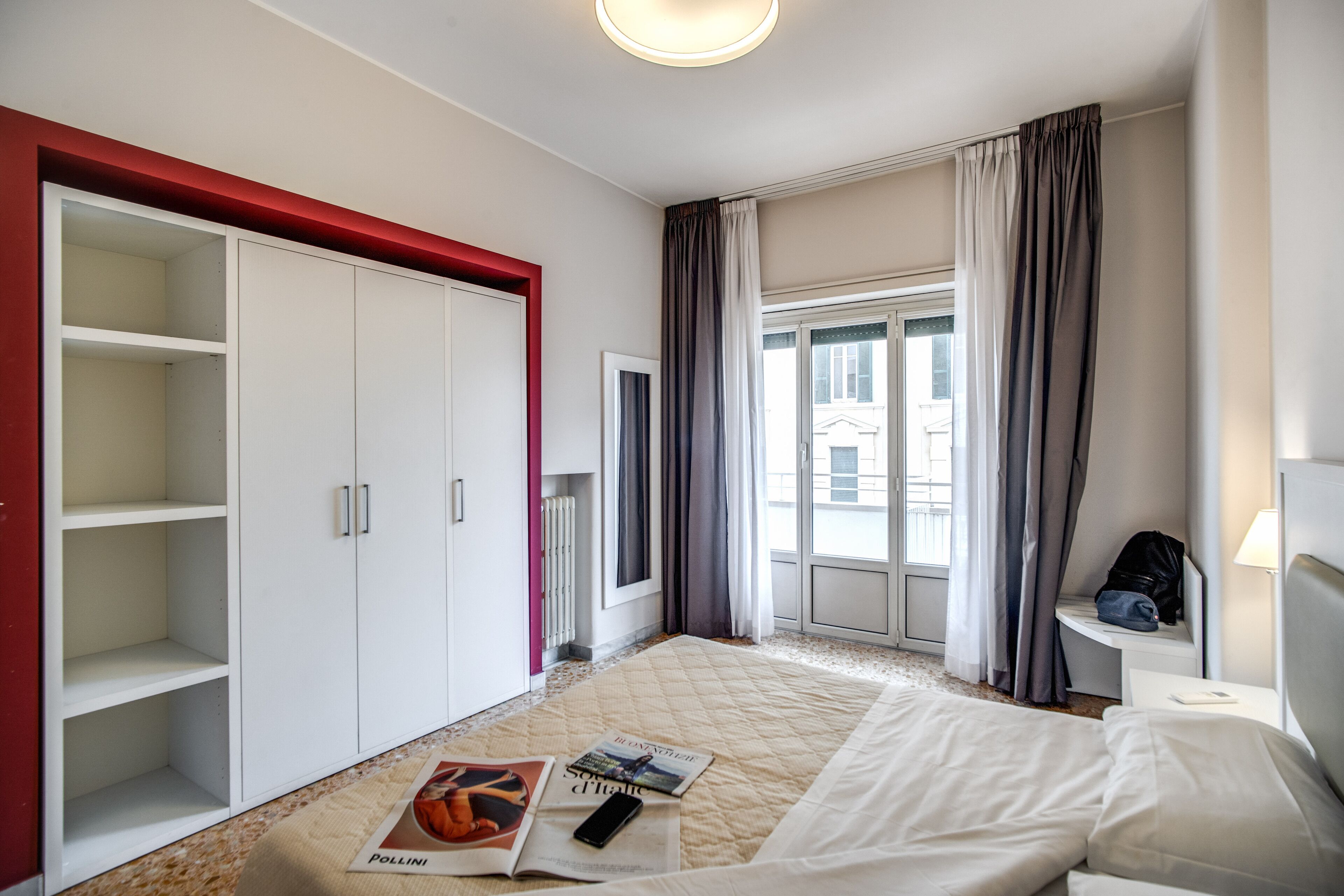 A hotel room with a bed, a wardrobe, a window with curtains, and a bedside table with a lamp.