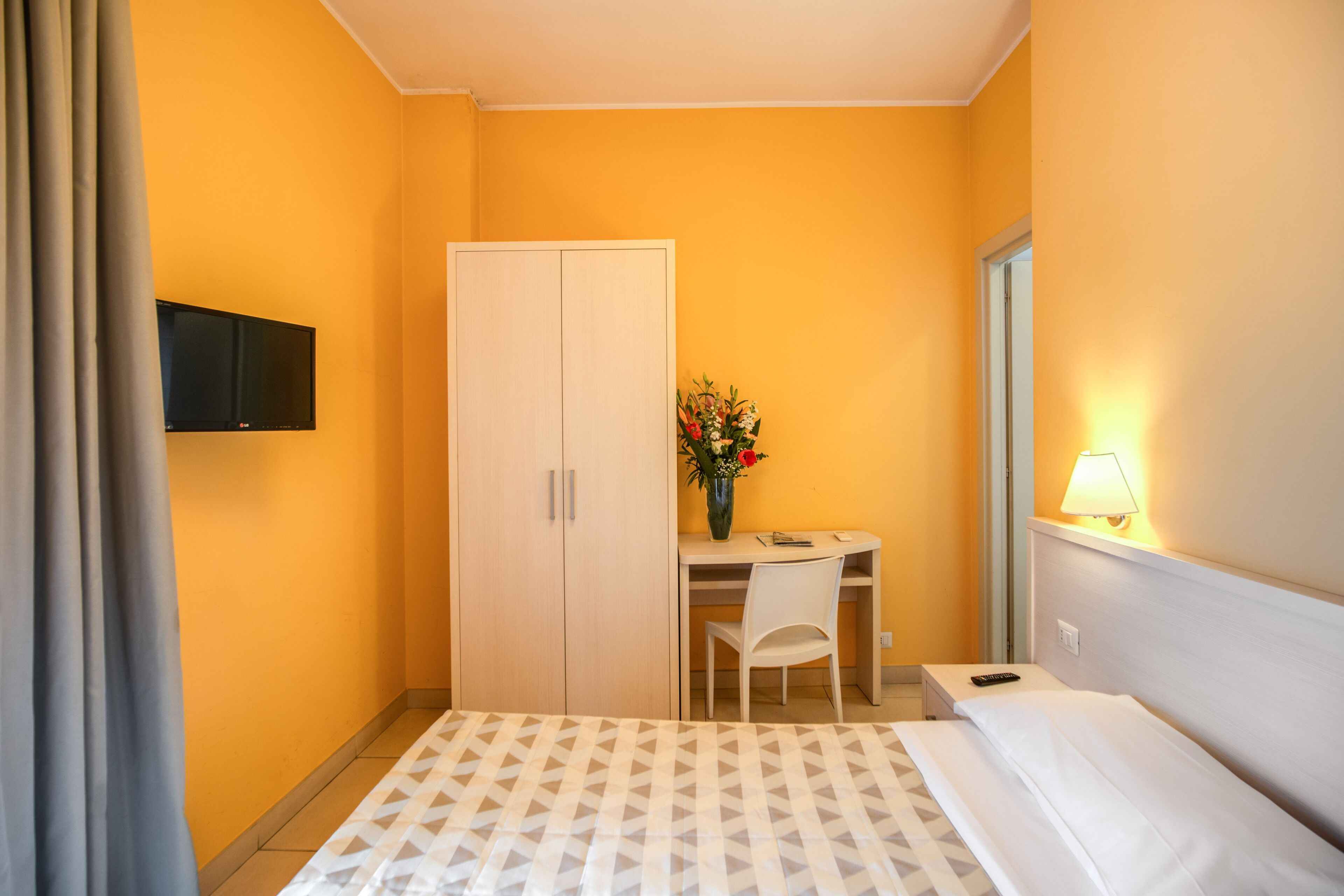 A hotel room with a bed, a desk with a chair, a wardrobe, a TV mounted on the wall, and a lamp on the bedside table.