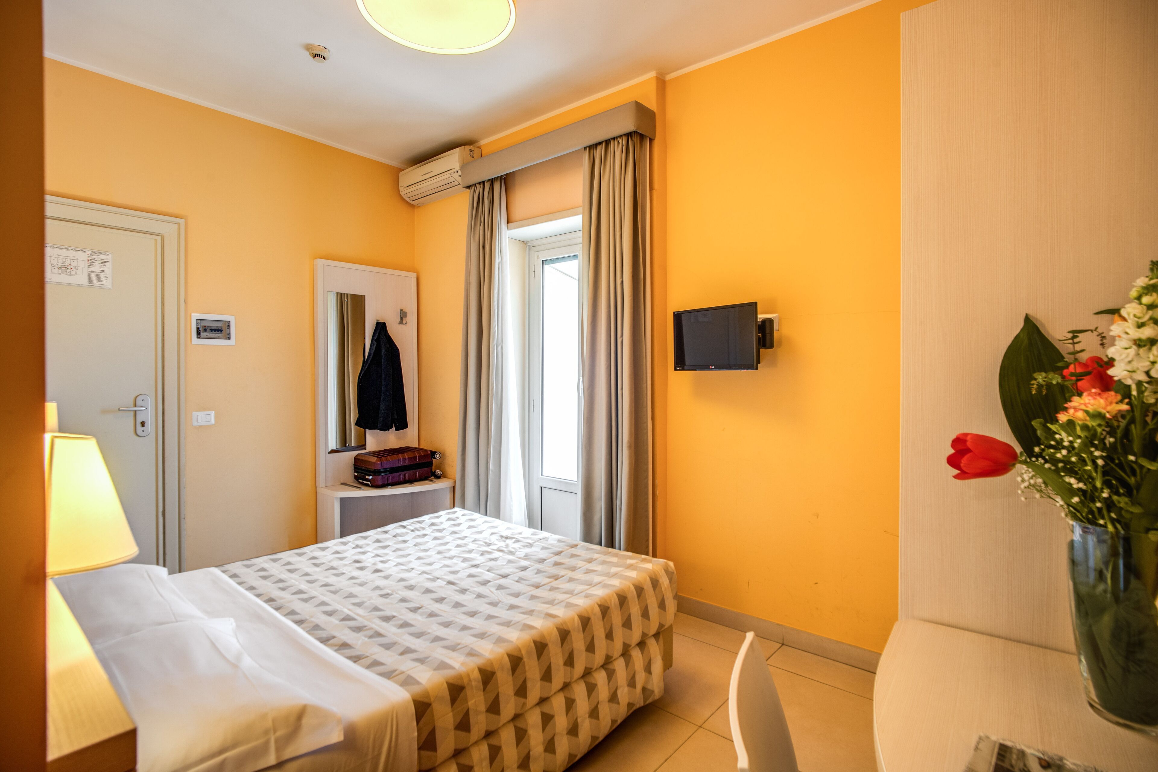 A hotel room with a bed, a desk, a chair, a TV, a window with curtains, a vase with flowers, and a door with a lock.