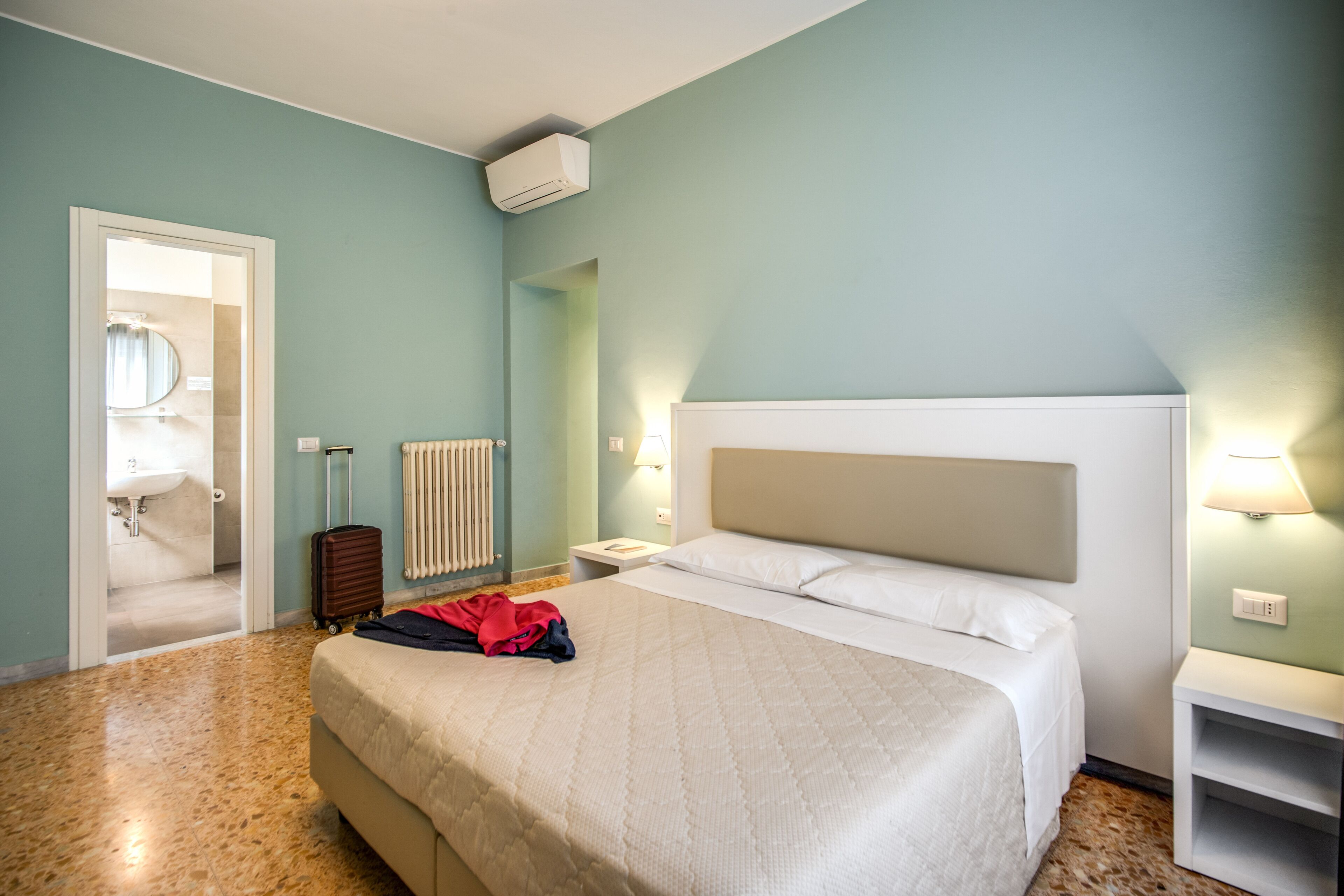 Hotellihuone, jossa on sänky, matkalaukku, patteri ja avoimesta ovesta näkyvä kylpyhuone.