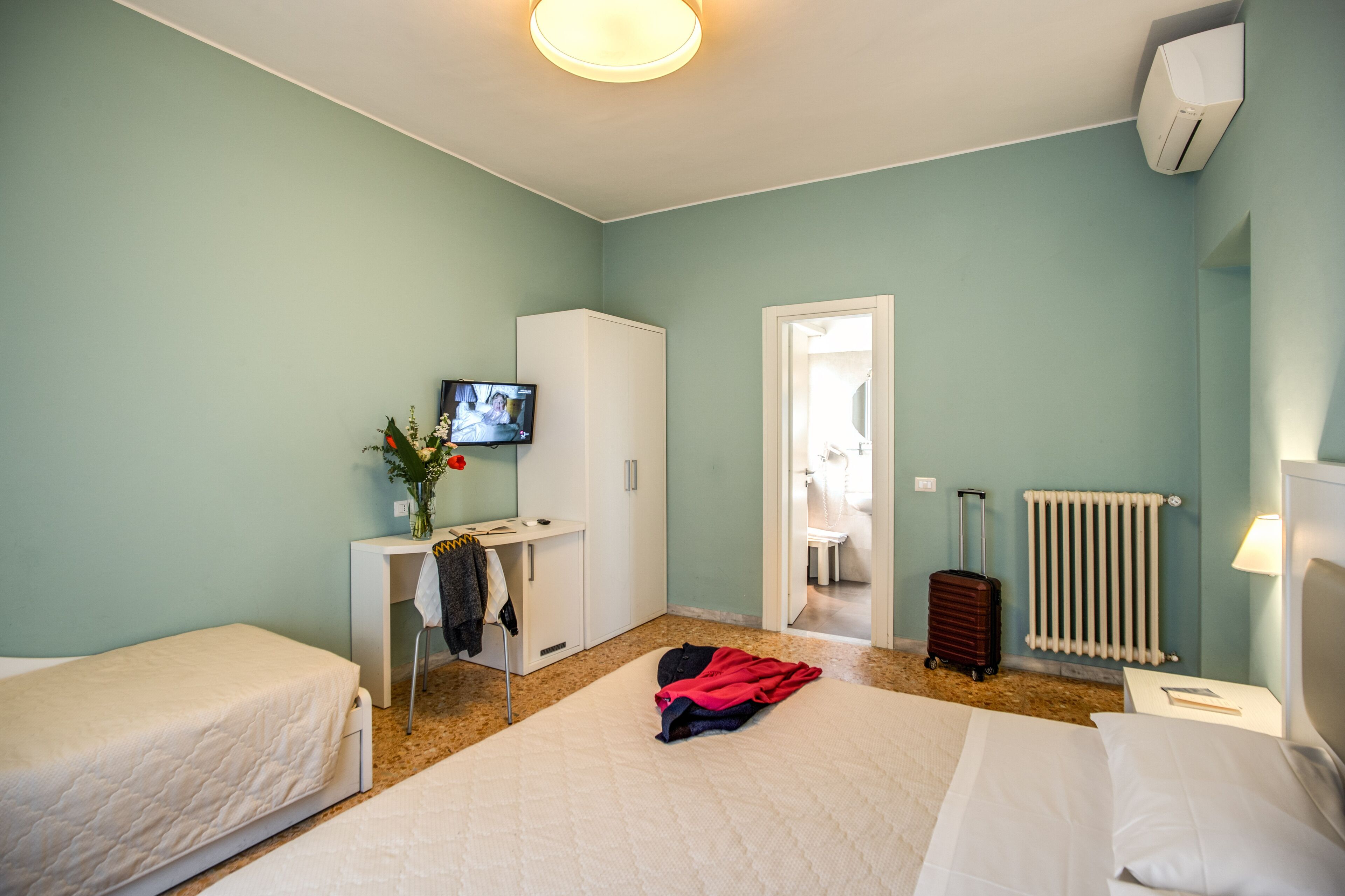 A hotel room with two beds, a desk with a TV, a radiator, and a suitcase.