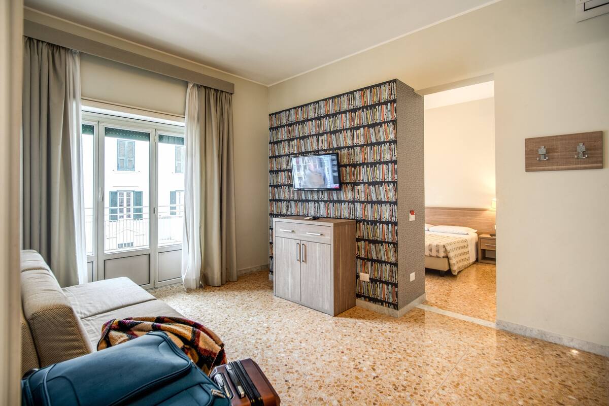 A hotel room with a sofa, a television mounted on a bookshelf, a bed, and a suitcase on the floor.