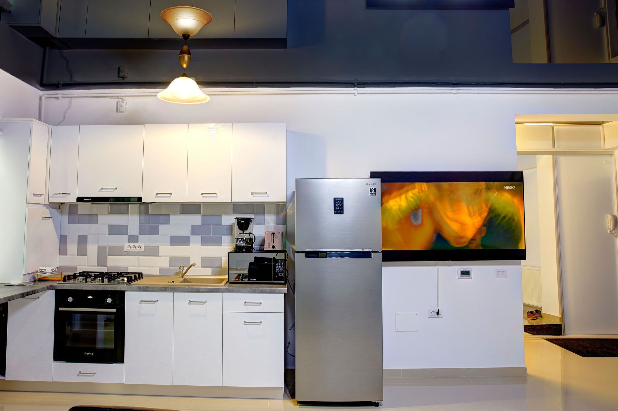 A modern kitchen with white cabinets, a stainless steel refrigerator, a built-in oven, and a flat-screen TV mounted above the counter.