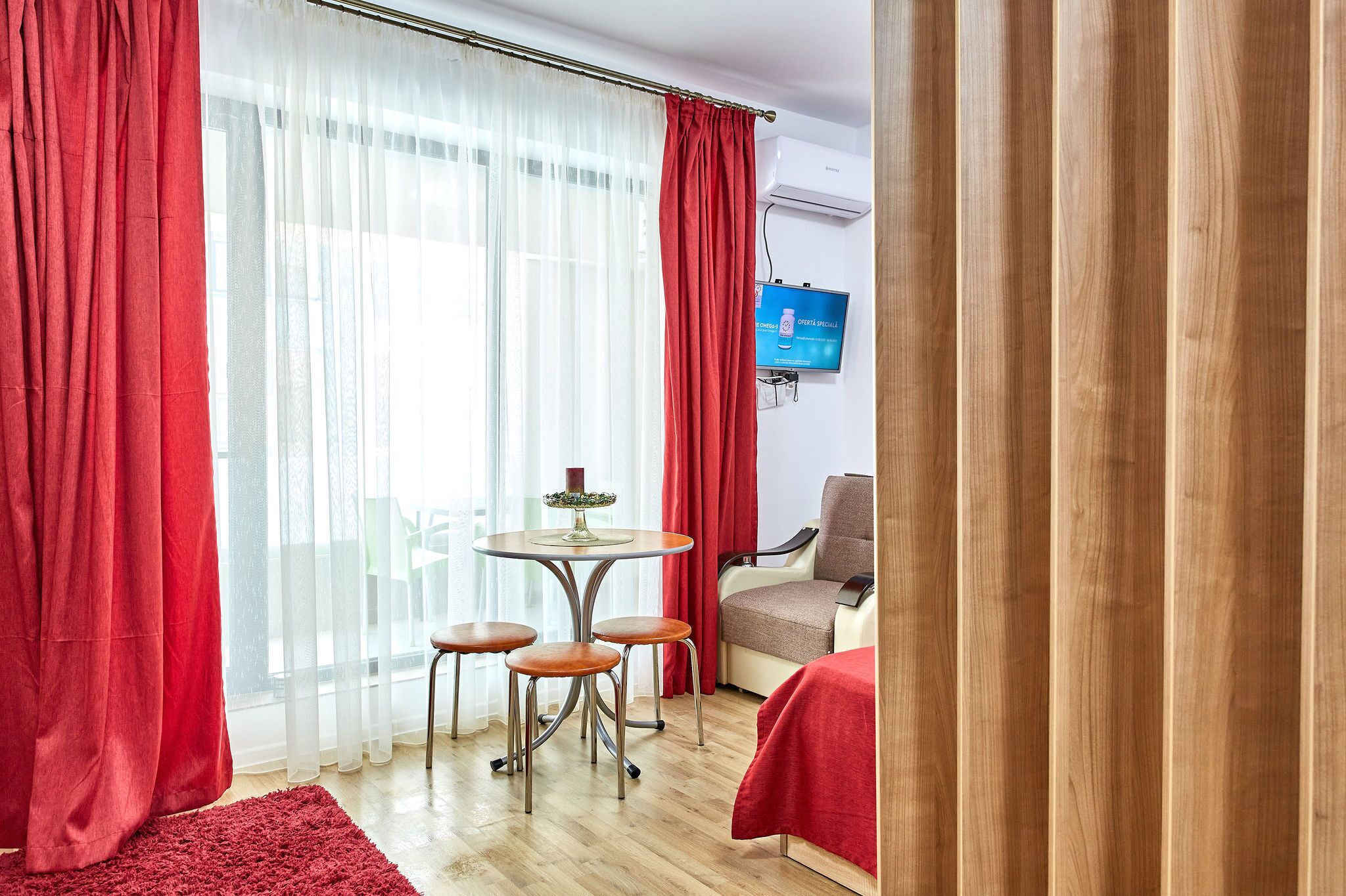 Habitación de hotel con una cama, un televisor montado en la pared, una mesita con sillas y un ventanal con cortinas de gasa y una cortina roja.
