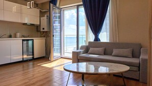 A modern living room with a sofa and a glass coffee table, a kitchen with white cabinets, and a large window with a view of the sea. - Apartamente Papaya (Navodari)