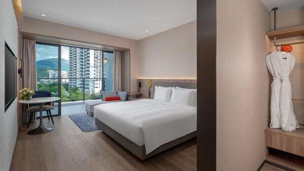 Habitación de hotel con una cama grande, un escritorio, una silla, una bata colgada en un perchero y vistas de la ciudad a través de la ventana.