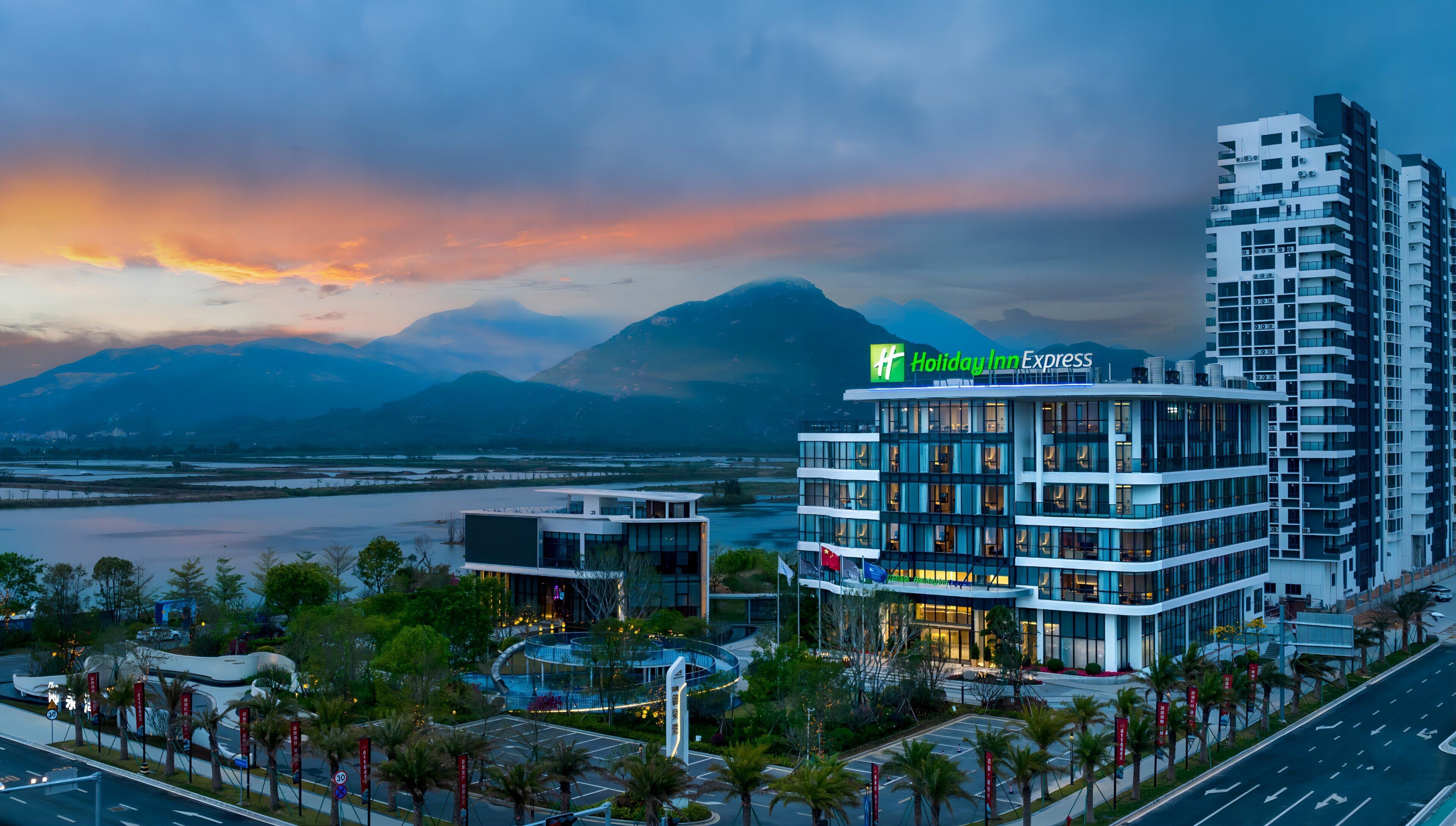 Un hôtel Holiday Inn Express au design moderne, situé près d’un plan d’eau, avec des montagnes en arrière-plan.
