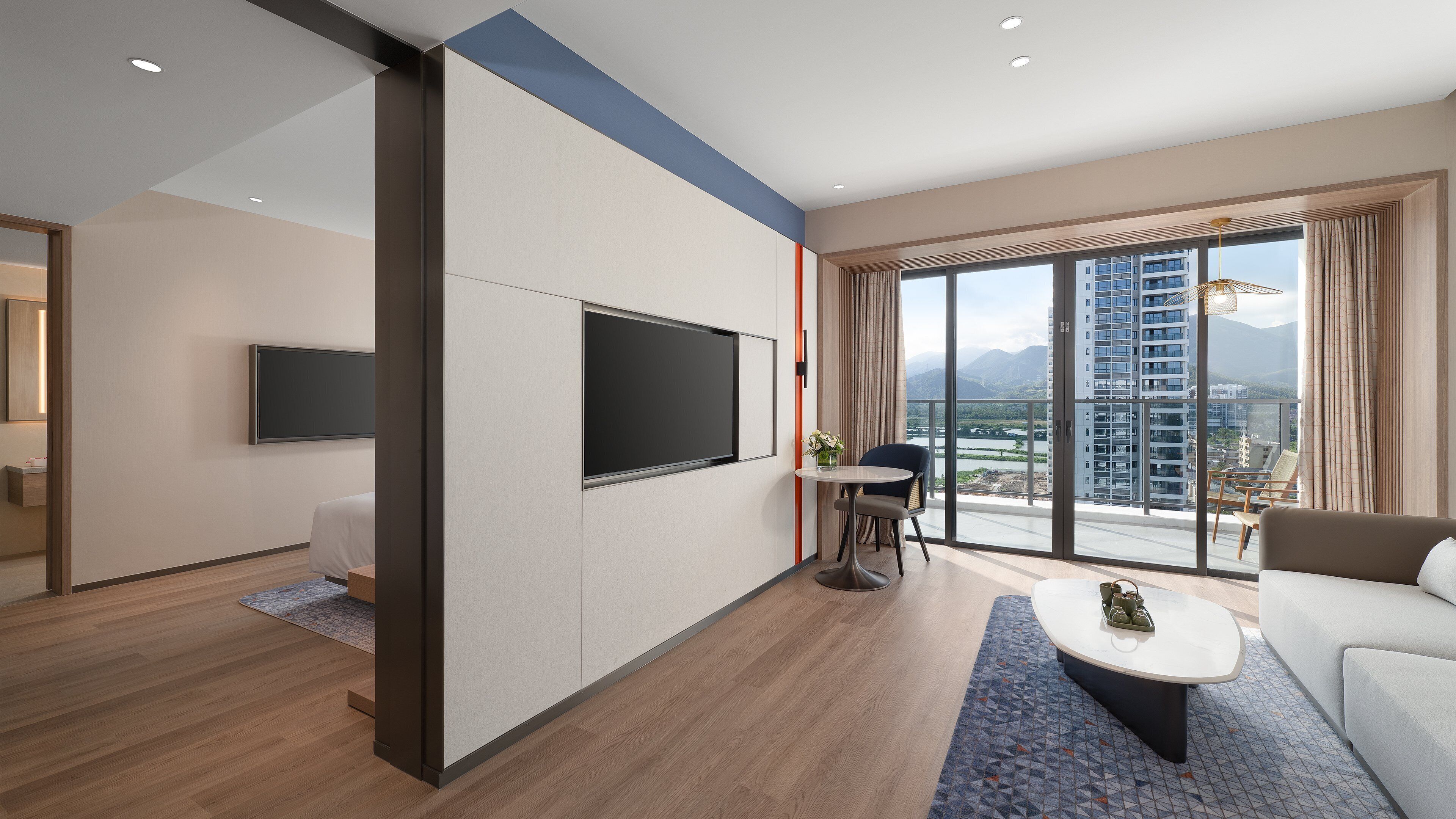 A modern hotel room with a large TV, a balcony with a city view, and a cozy seating area.