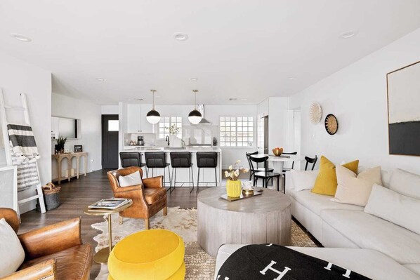 A modern living room with a sofa, ottoman, and chairs, a dining area with a table and chairs, and a kitchen with bar stools.