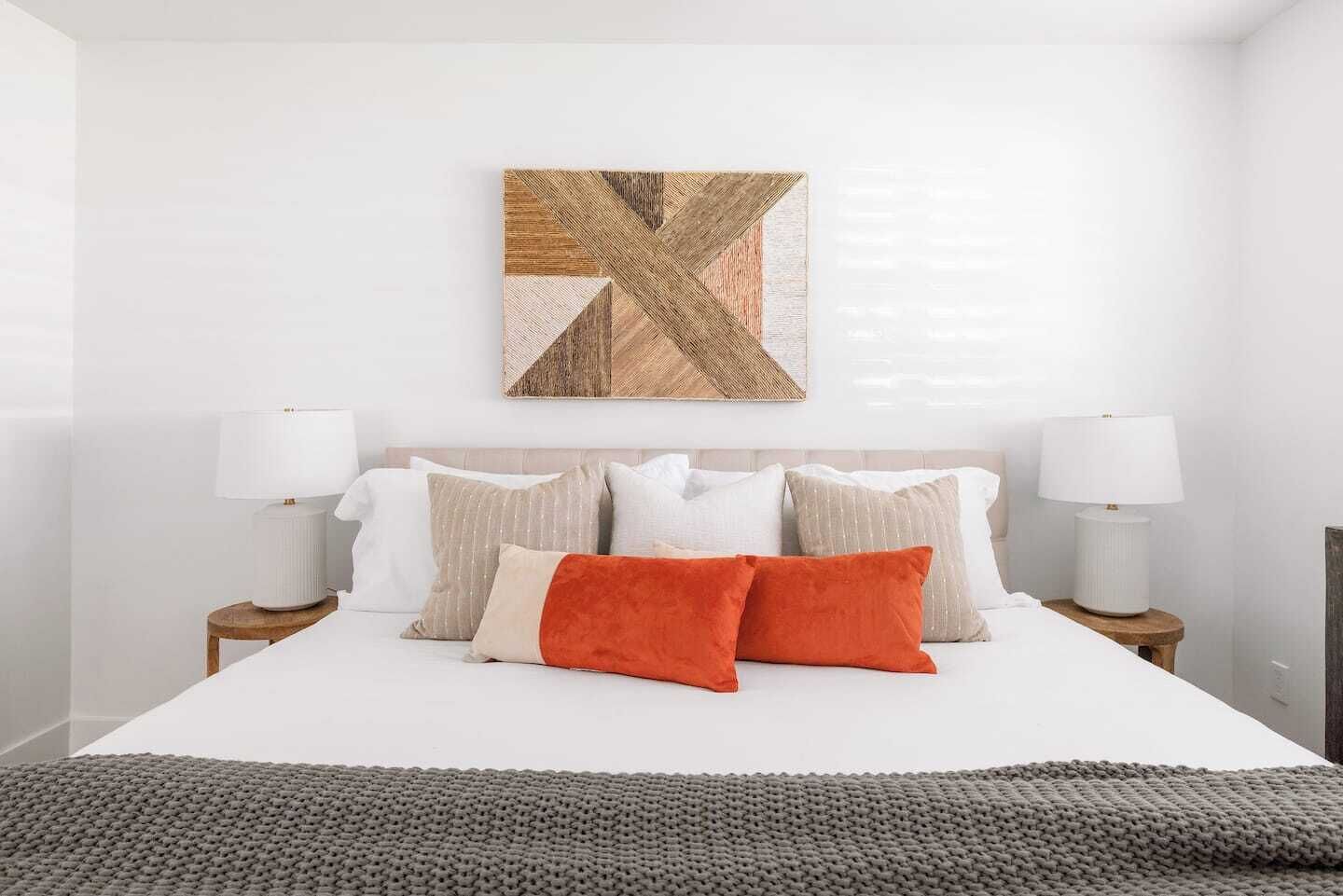 Un dormitorio ordenado con una cama grande, dos mesitas de noche con lámparas, y una obra de arte geométrica en la pared.
