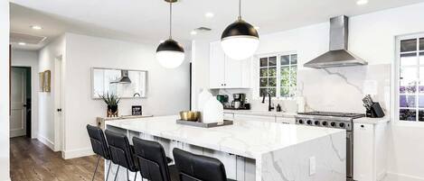 A modern kitchen with a large island, black bar stools, pendant lights, and a stainless steel range hood.