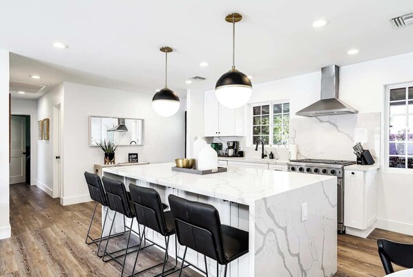 A modern kitchen with a large island, black bar stools, pendant lights, and a stainless steel range hood.