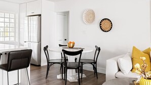 Une cuisine moderne avec des meubles blancs, un coin repas doté d’une table ronde et de chaises, et un salon avec un canapé et des coussins.