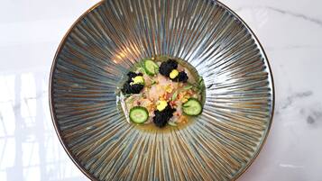 Uma placa metálica redonda com padrão radial, contendo um prato com caviar preto, fatias de pepino e um guarnição de ervas e nozes.