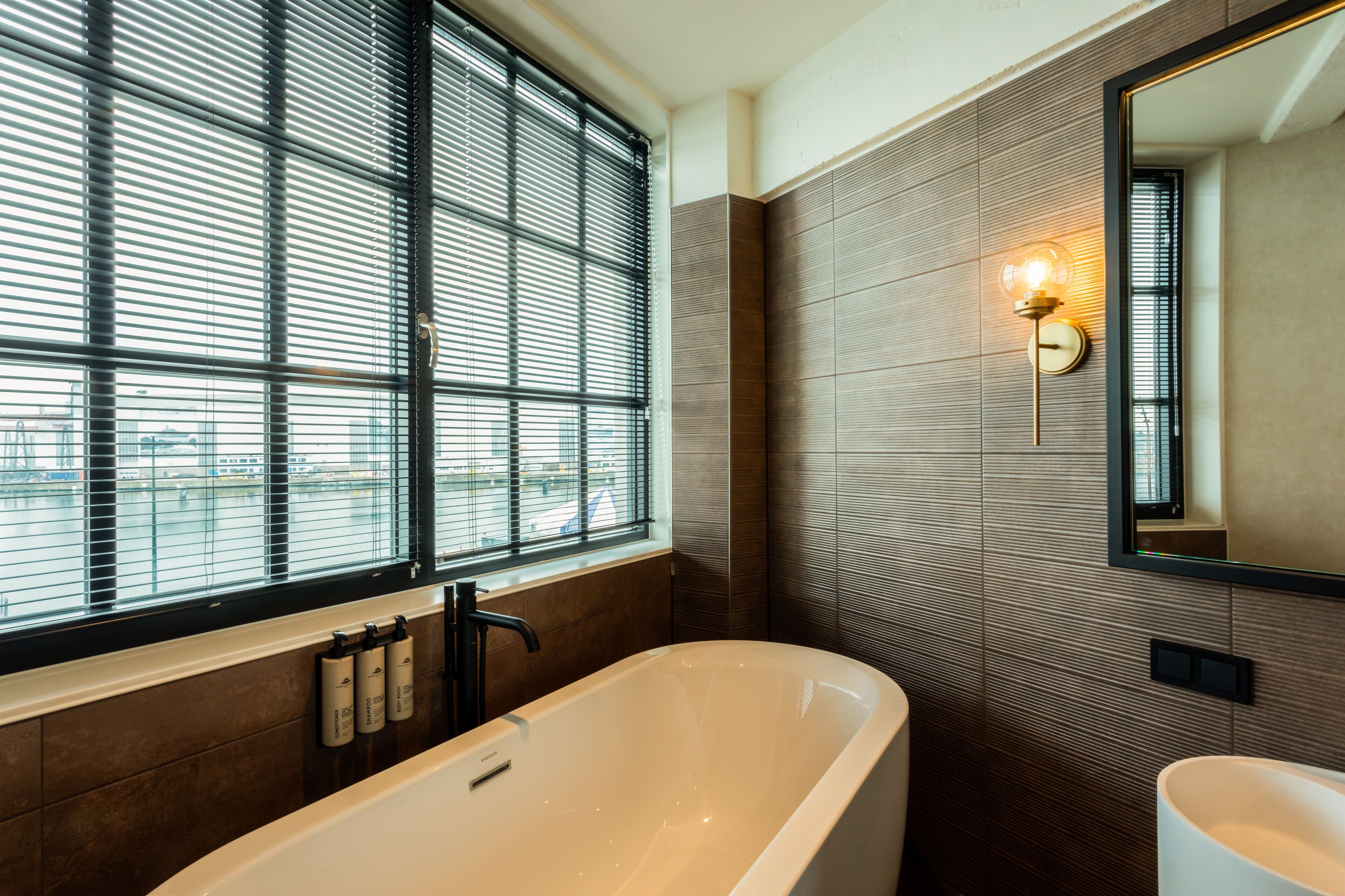 A modern bathroom with a freestanding bathtub, a large window with blinds, and a wall-mounted mirror.