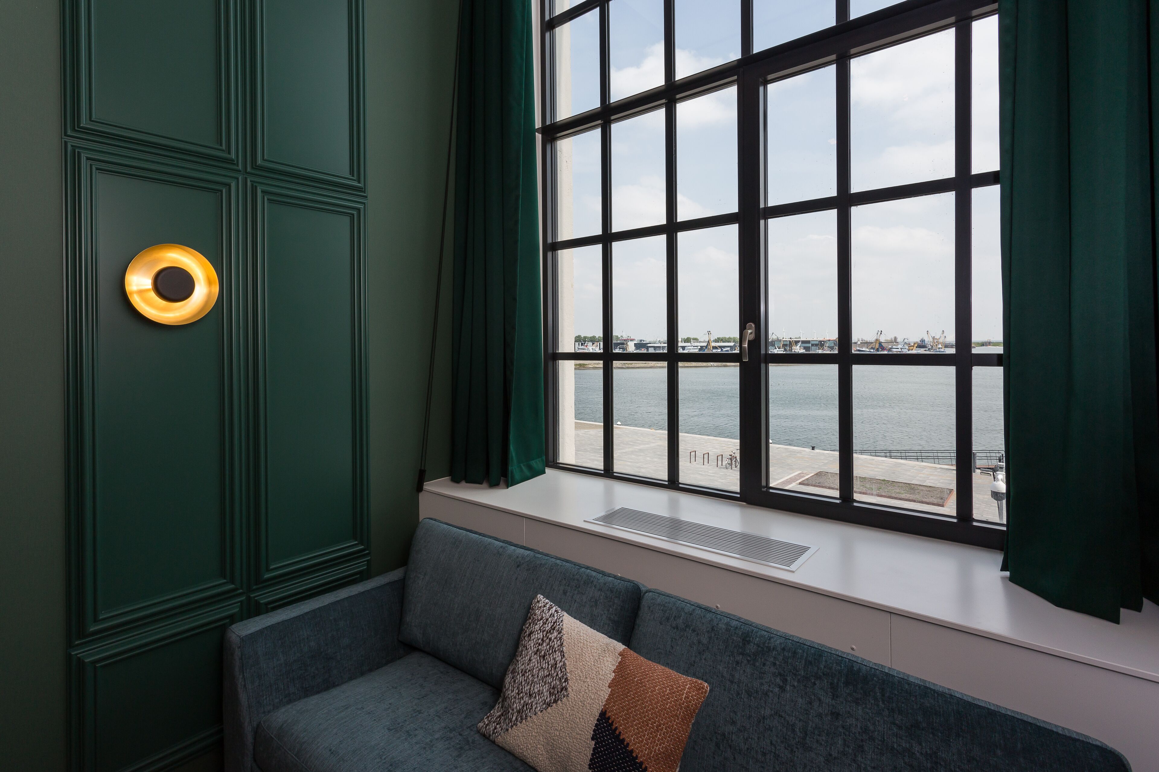 A room with a large window overlooking a body of water, a green wall, and a grey sofa with a patterned pillow.