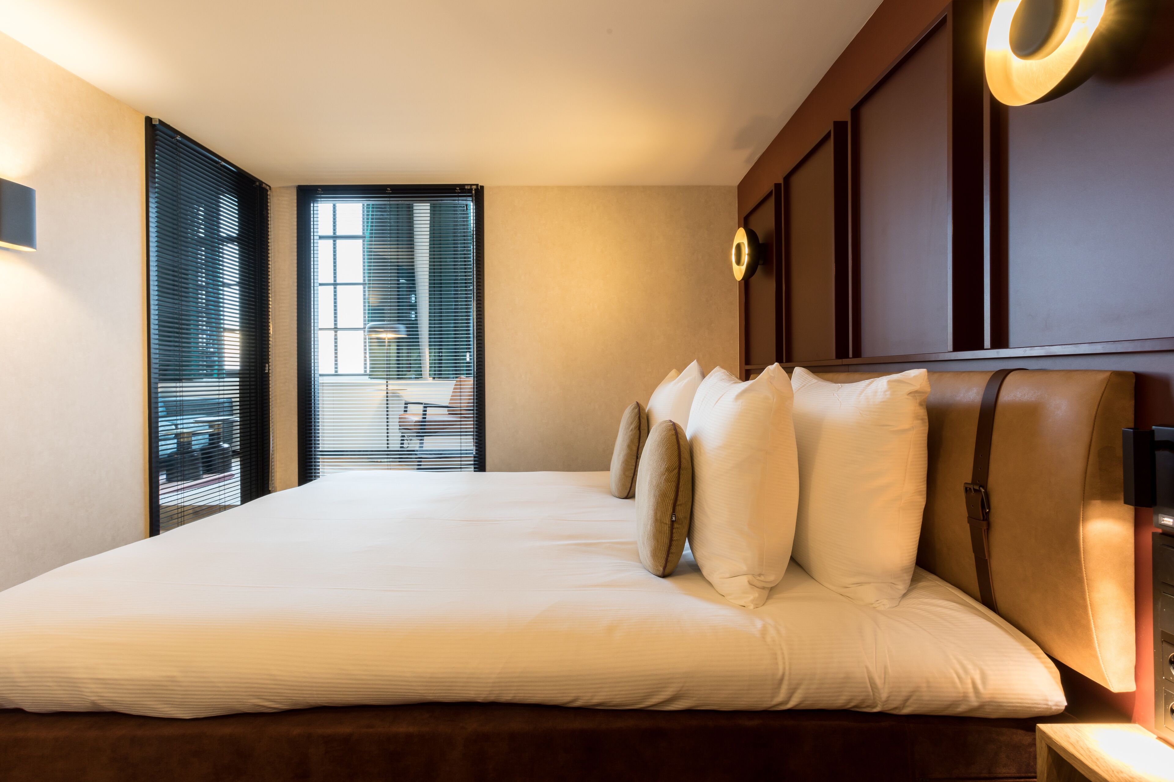 Une chambre d’hôtel avec un grand lit, une tête de lit dotée d’un éclairage circulaire et une vue à travers les stores de la fenêtre.