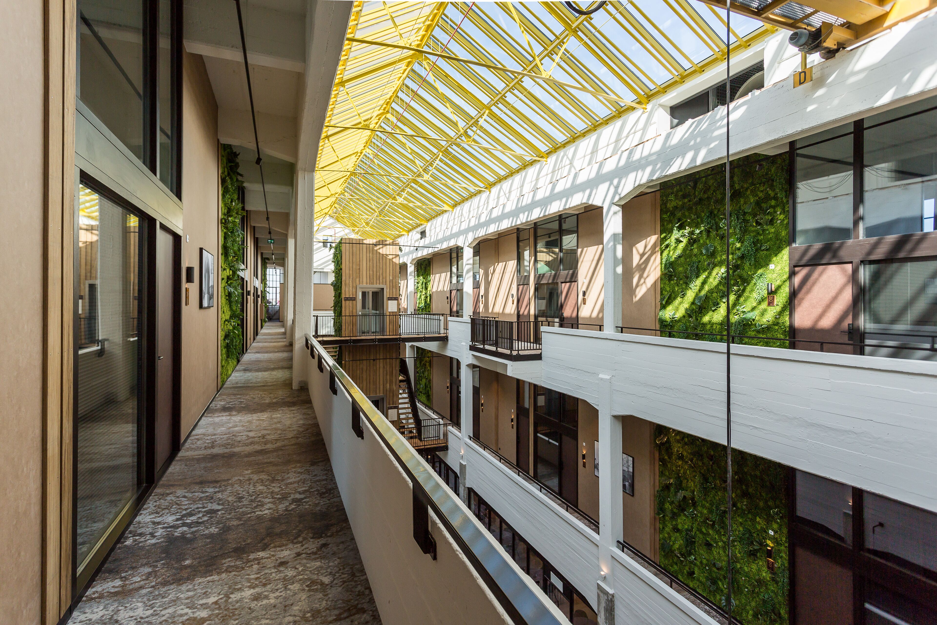 A modern hotel corridor with a glass ceiling, indoor plants, and multiple balconies.