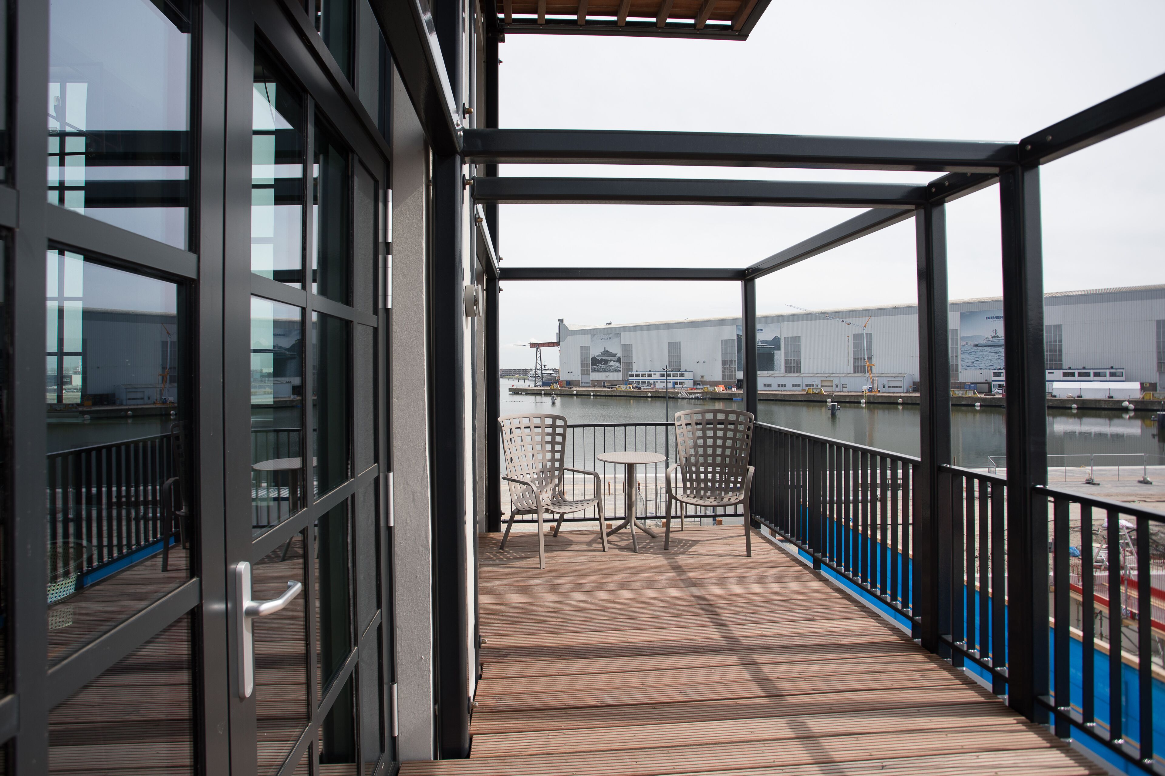 A covered balcony with a view of a large industrial building and a body of water.