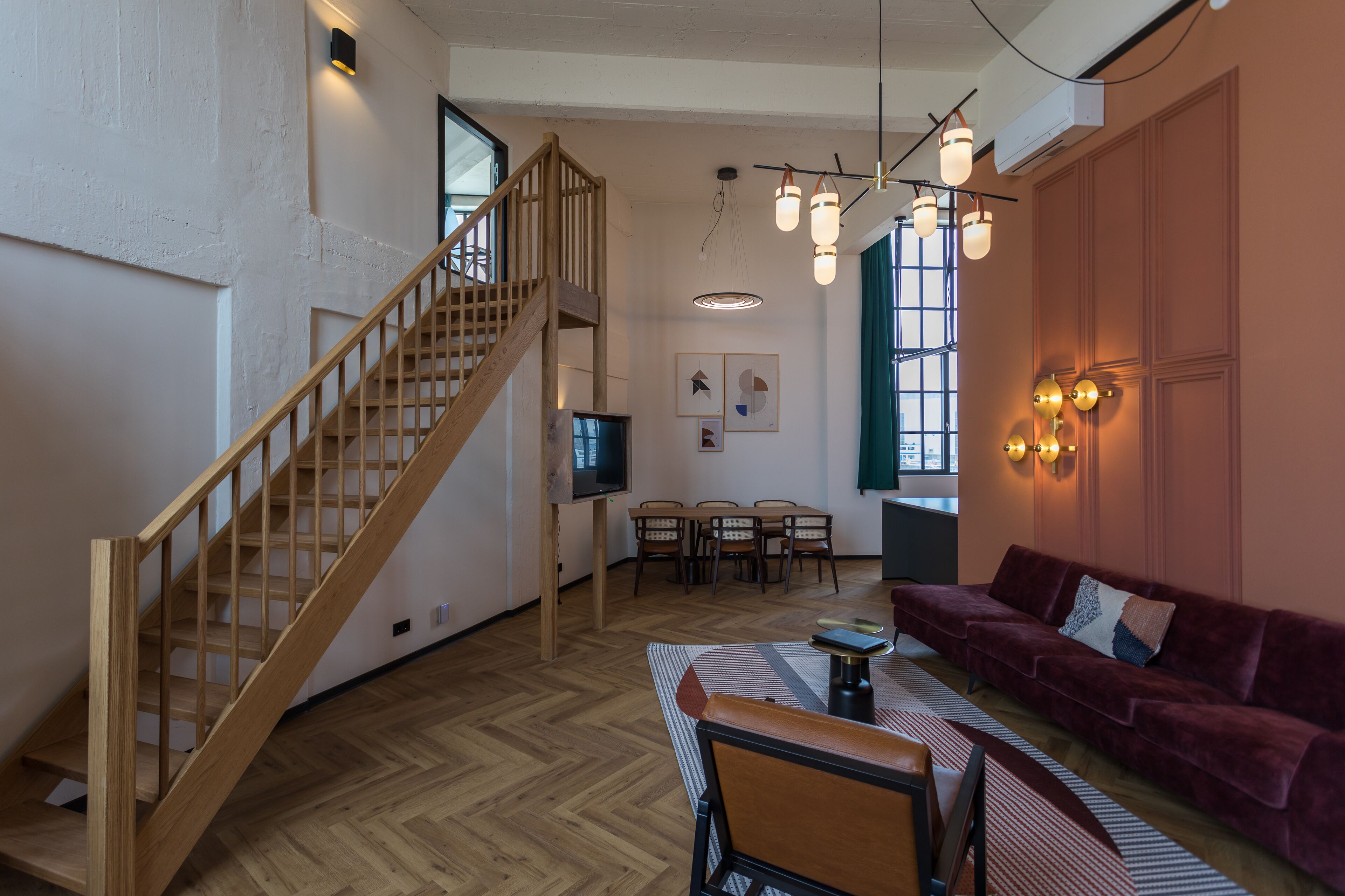 A spacious living area with a wooden staircase, a dining table with chairs, a sofa, and a television.