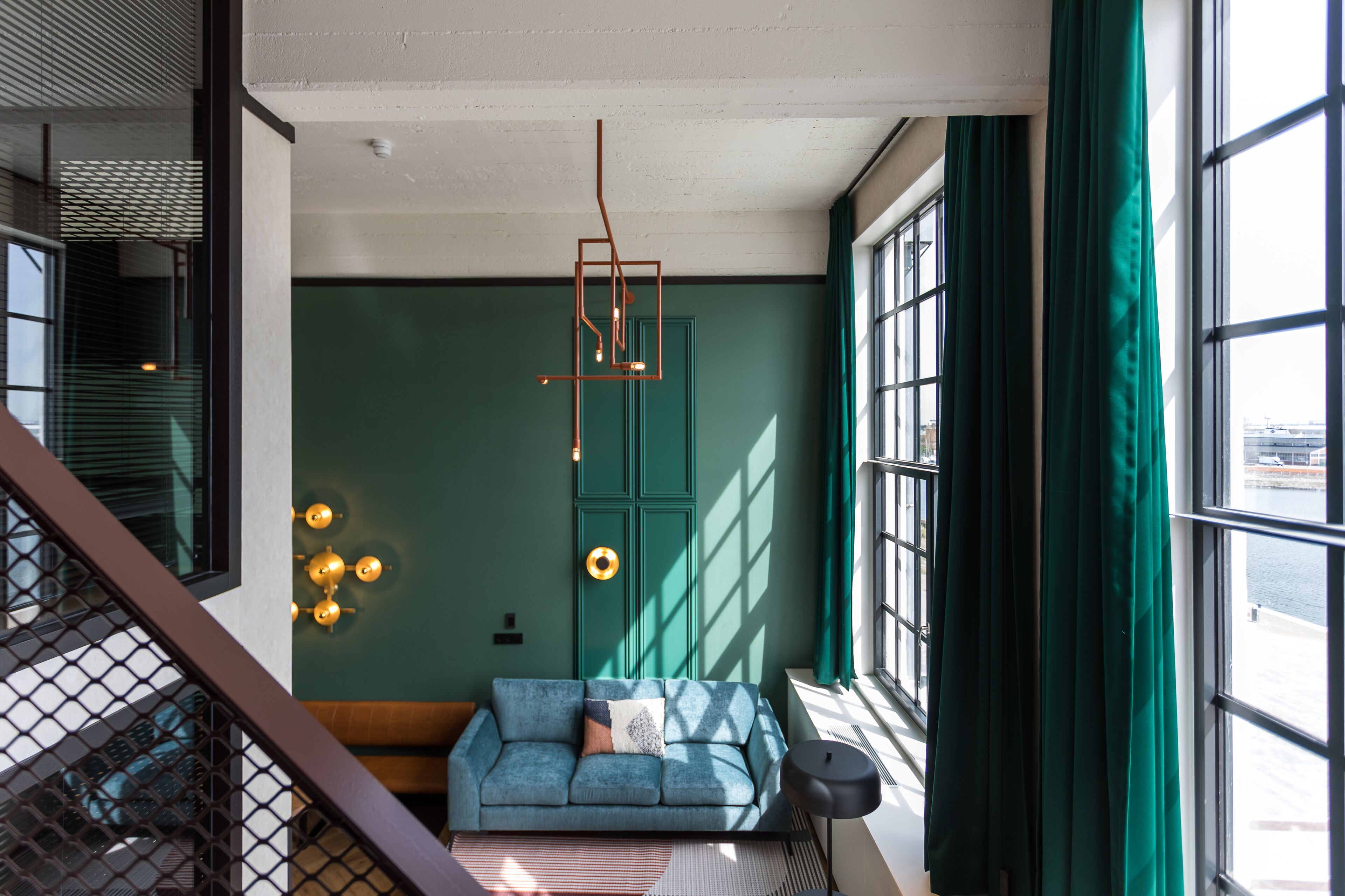 Een moderne woonkamer met een blauwgroene bank, een hanglamp en grote ramen met groene gordijnen.