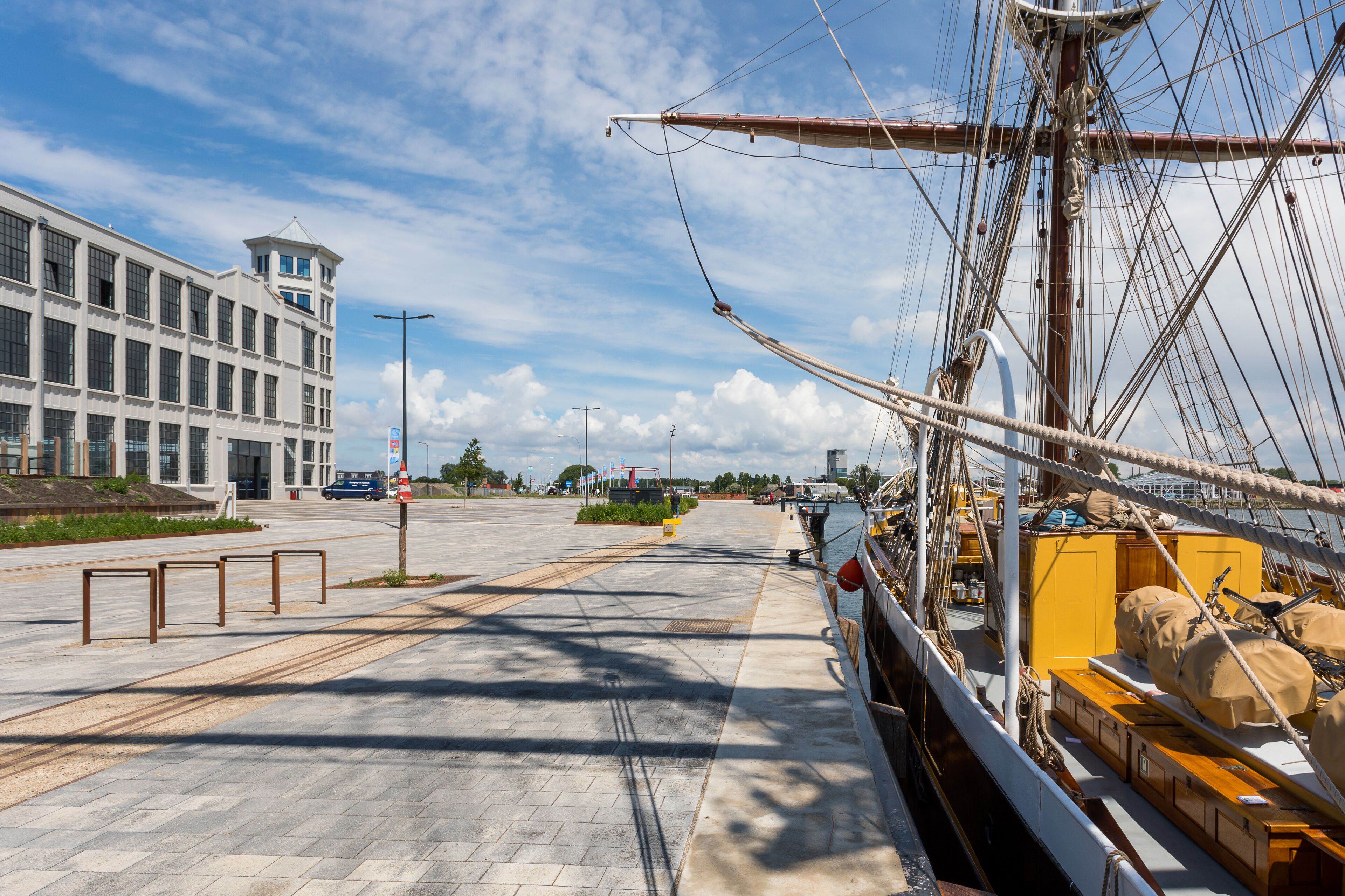 En promenade langs vandet med en moderne bygning og et sejlskib i nærheden.