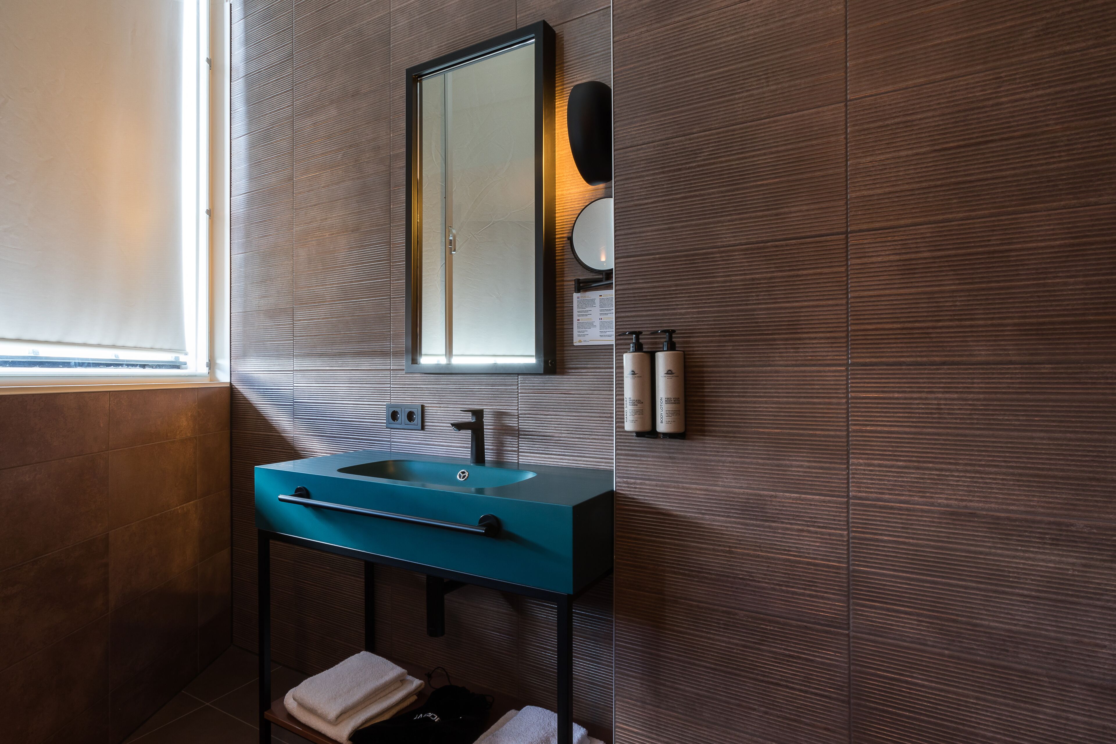 A modern bathroom with a teal sink, a large mirror, and a wall-mounted soap dispenser.