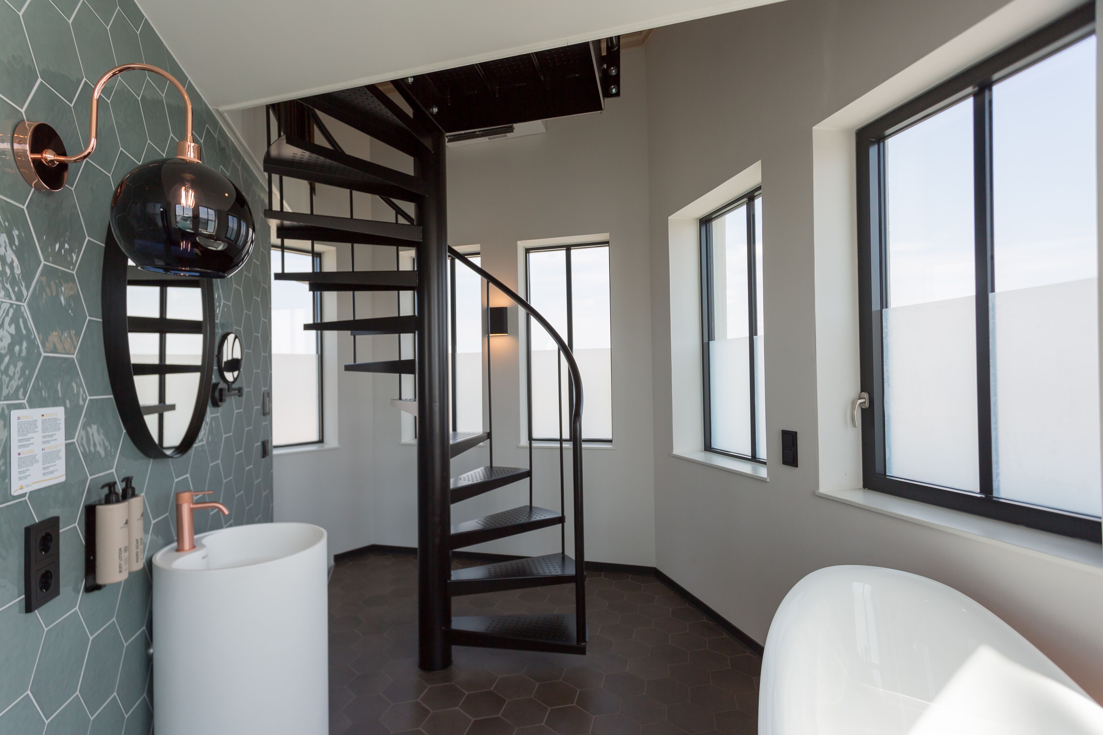 Un baño moderno con un lavamanos grande blanco, una escalera en espiral negra y un amplio ventanal que proporciona luz natural.