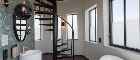 Un baño moderno con un lavamanos grande blanco, una escalera en espiral negra y un amplio ventanal que proporciona luz natural.