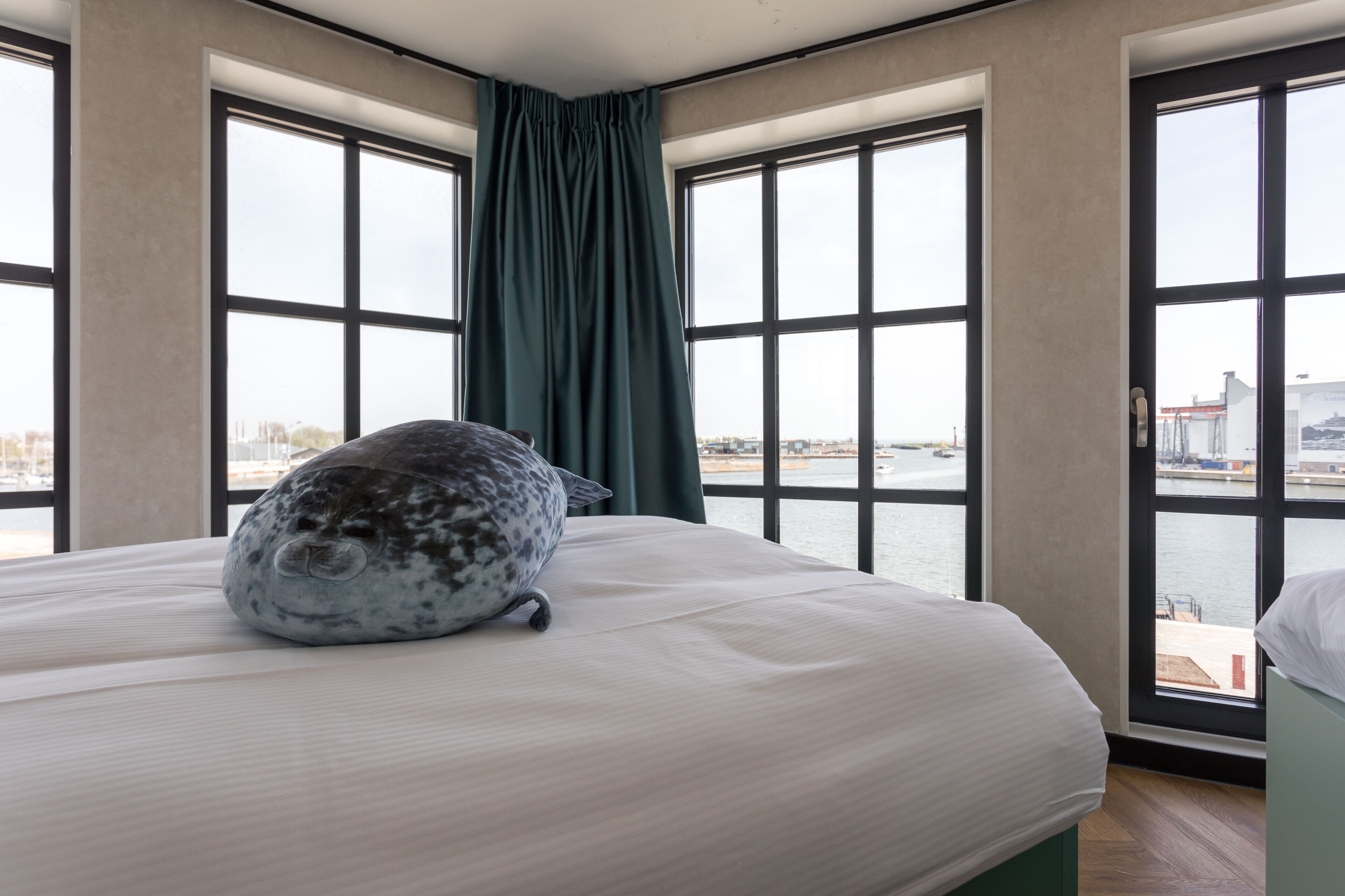 Une chambre avec un lit, une grande fenêtre donnant sur un plan d’eau et sur le paysage urbain, et un rideau.