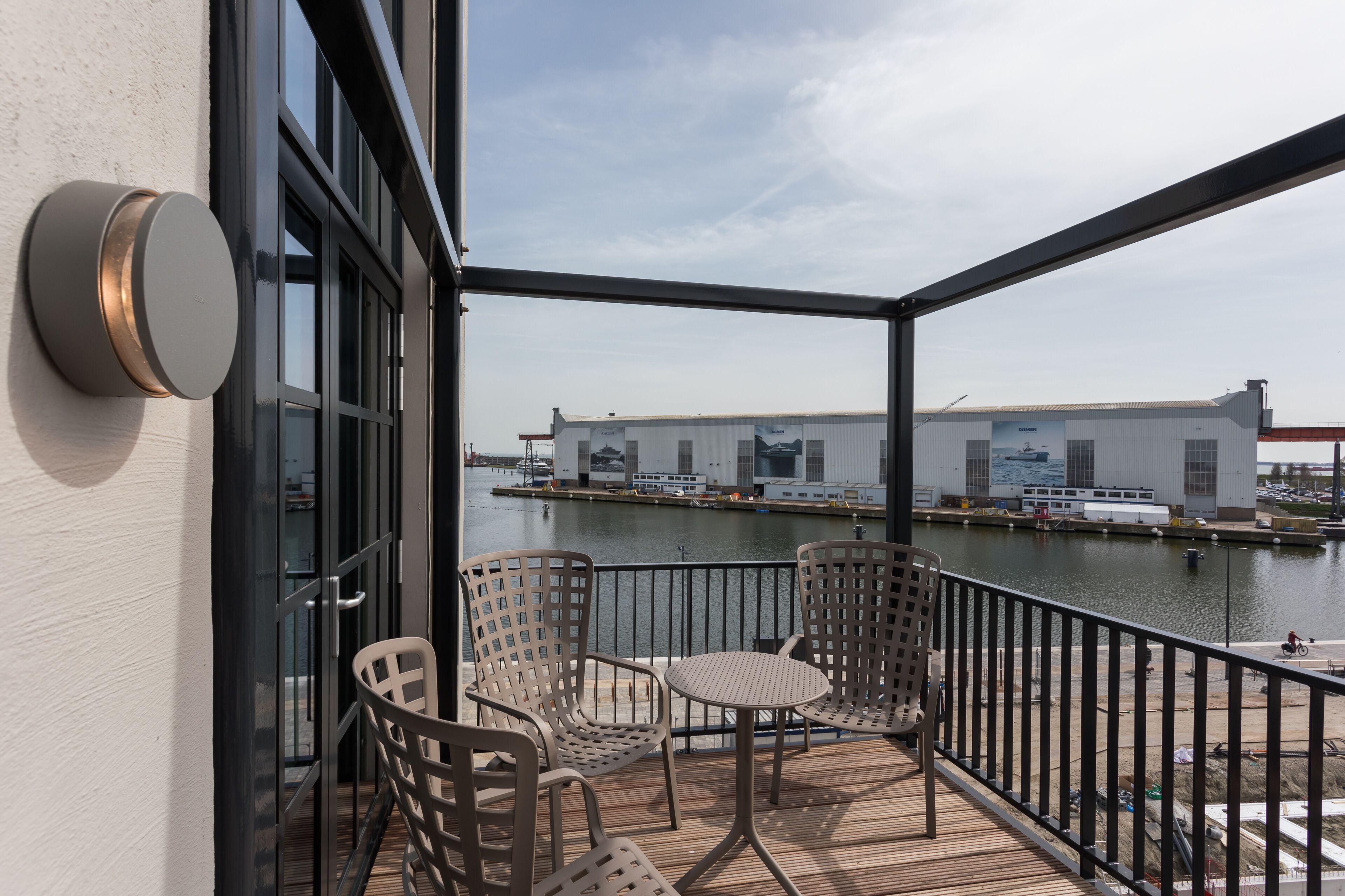 A balcony with wicker chairs and a small table, overlooking a body of water and a large industrial building.