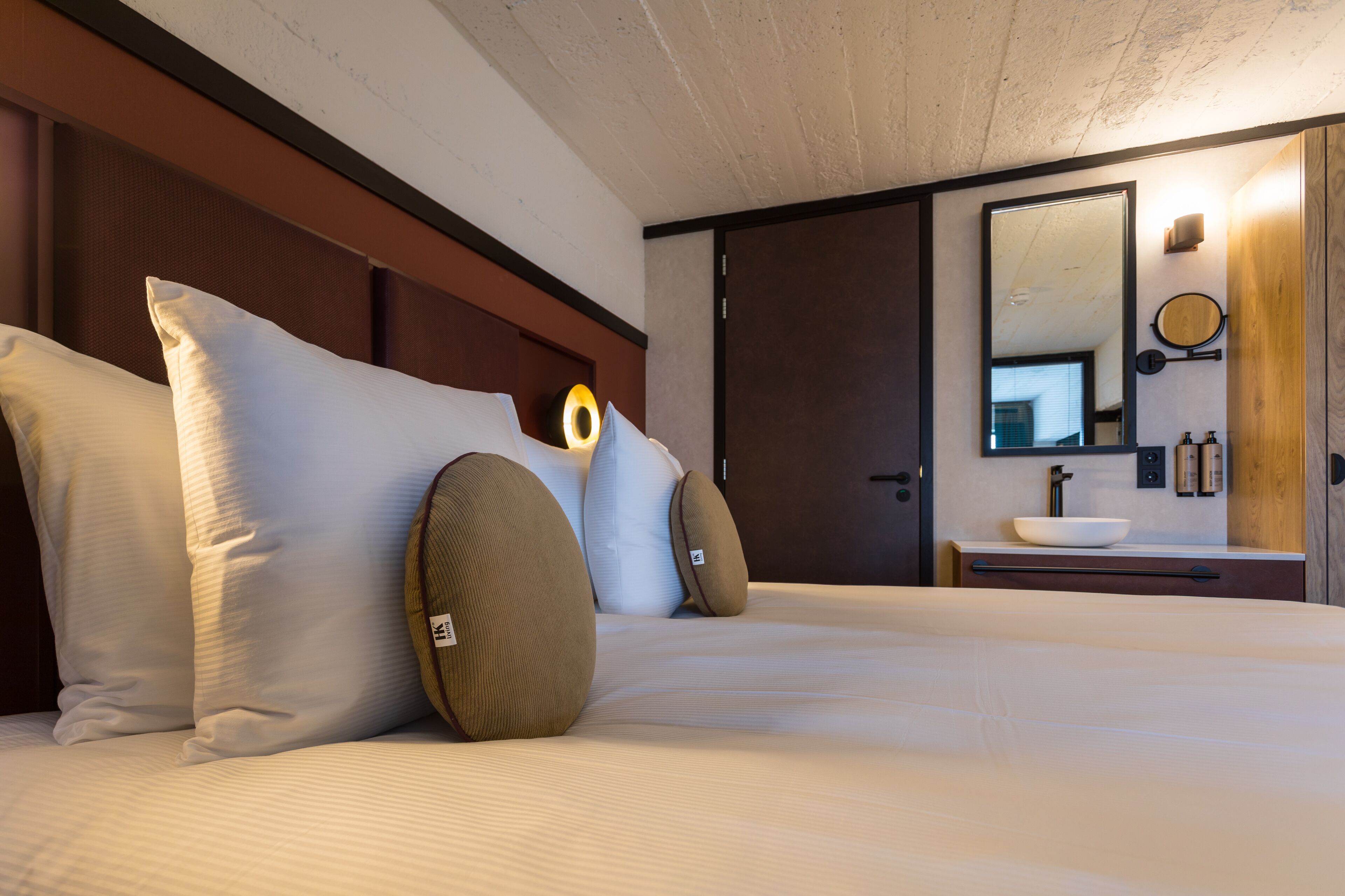 A neatly made bed with pillows, a bedside table, a mirror, and a small sink with toiletries.