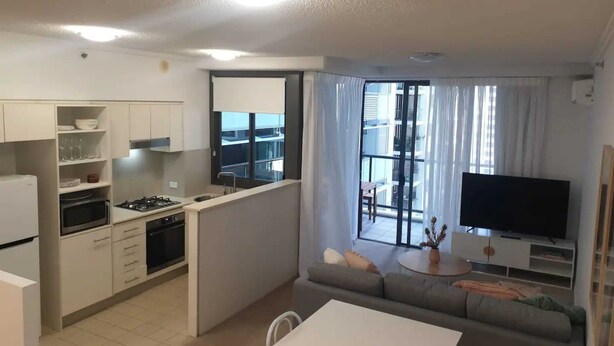 A modern apartment with a kitchen, dining area, and living room. It includes a large TV, a sofa, and a balcony with a view. - Open 1 Bedroom Apartment in Brisbane City (Brisbane)