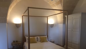 Una cama con dosel de madera con ropa de cama blanca, flanqueada por dos mesitas de noche con lámparas, en una habitación con arcos de piedra y un armario.
