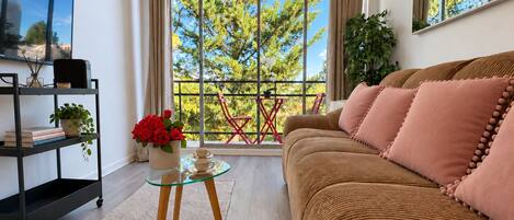 A modern living room with a sofa, a glass coffee table, a bookshelf, and a balcony with a view of trees.