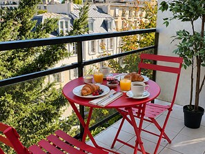 A red table with breakfast items, a chair, and a potted plant on a balcony with a city view.