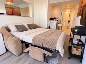Une chambre moderne avec un canapé-lit convertible, une table de chevet agrémentée d’un vase de fleurs et un petit bureau avec une chaise.