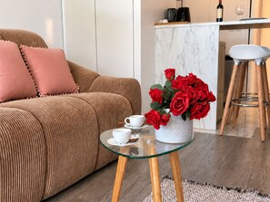 Un salon moderne avec un canapé, une table basse en verre, un vase de roses rouges et, au fond, un coin cuisine.