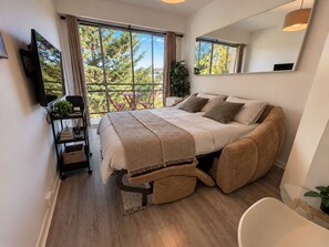 A modern bedroom with a large bed, a small desk, a chair, and a balcony with a view of trees.