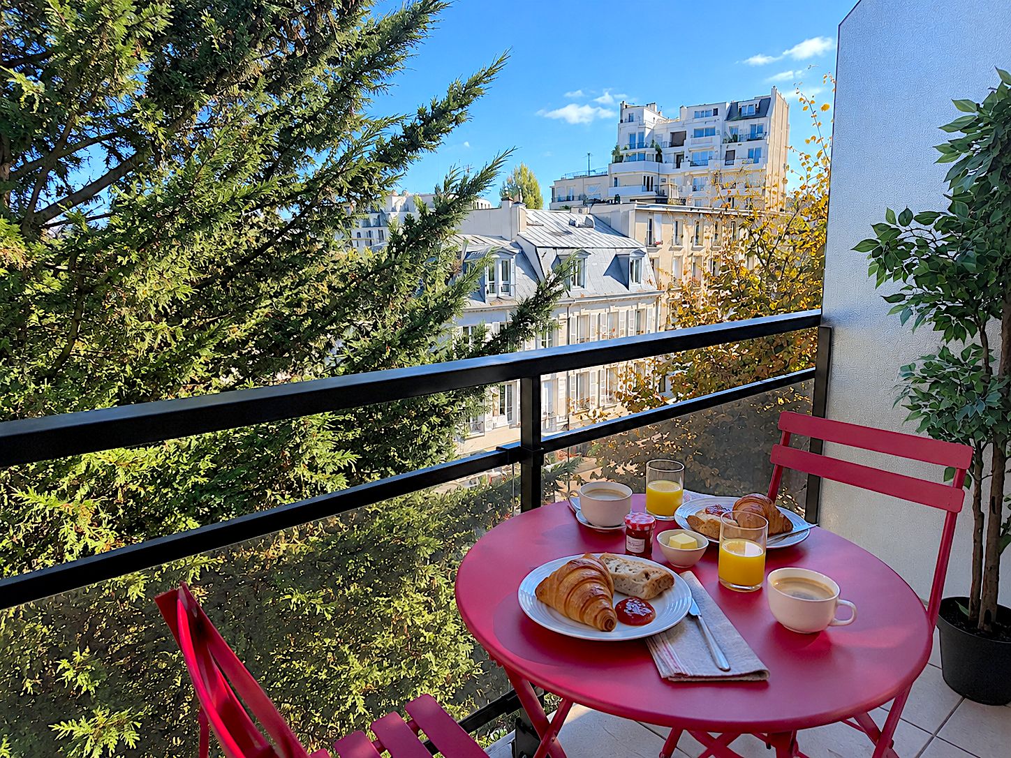 A balcony with a red table set for breakfast, including croissants, juice, and coffee, overlooking a cityscape.