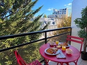 A balcony with a red table set for breakfast, including croissants, juice, and coffee, overlooking a cityscape.