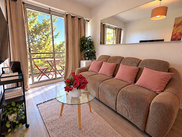 A modern living room with a sofa, a glass coffee table, and a view of a balcony with red chairs.